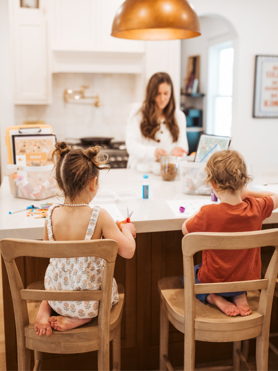 Two children sitting at a kitchen counter with a woman in the background.