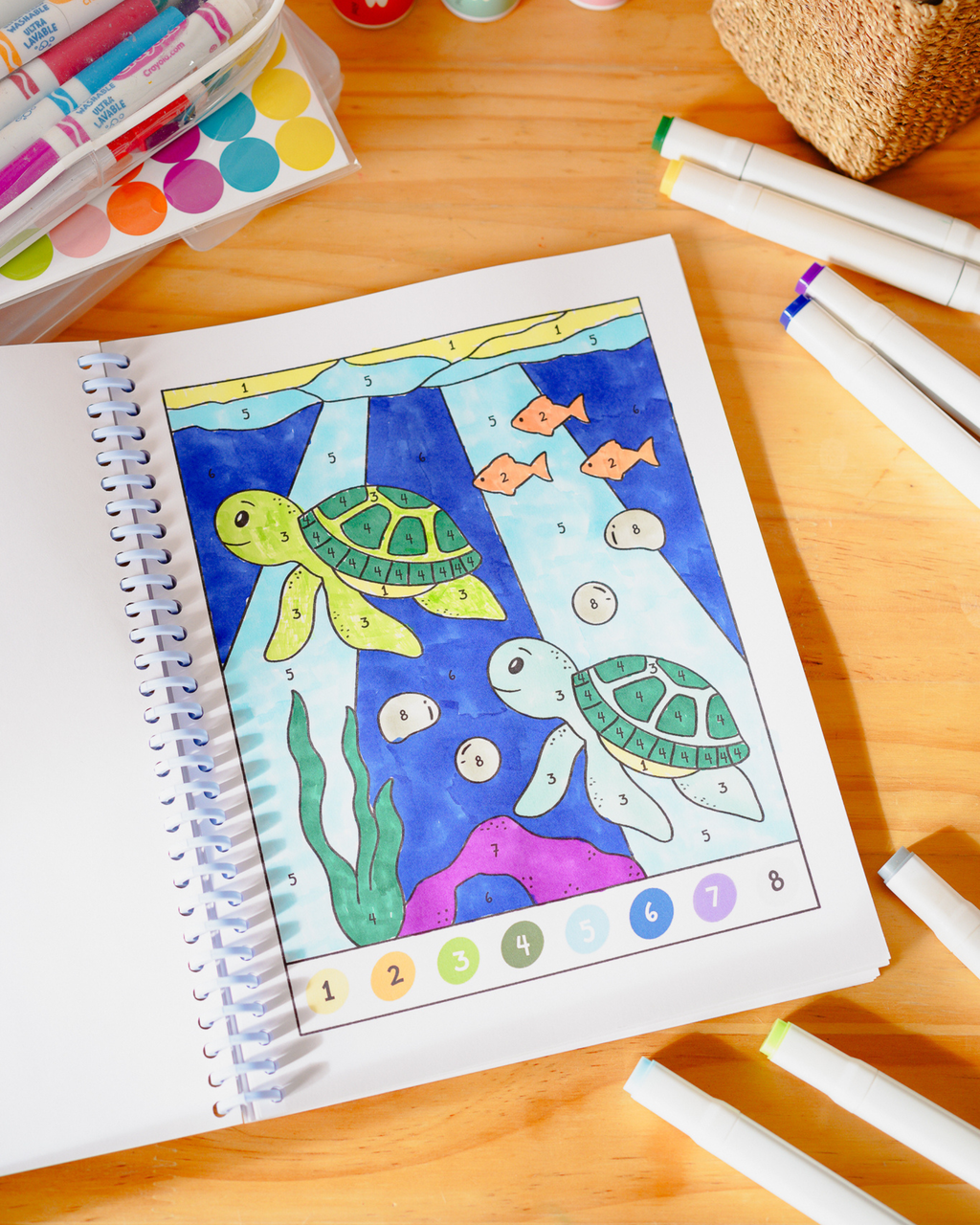 A children's coloring book or activity sheet on a wooden table, featuring an underwater scene with two sea turtles, fish, and numbered sections. Color markers and paint supplies are arranged around it.