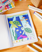A children's coloring book or activity sheet on a wooden table, featuring an underwater scene with two sea turtles, fish, and numbered sections. Color markers and paint supplies are arranged around it.