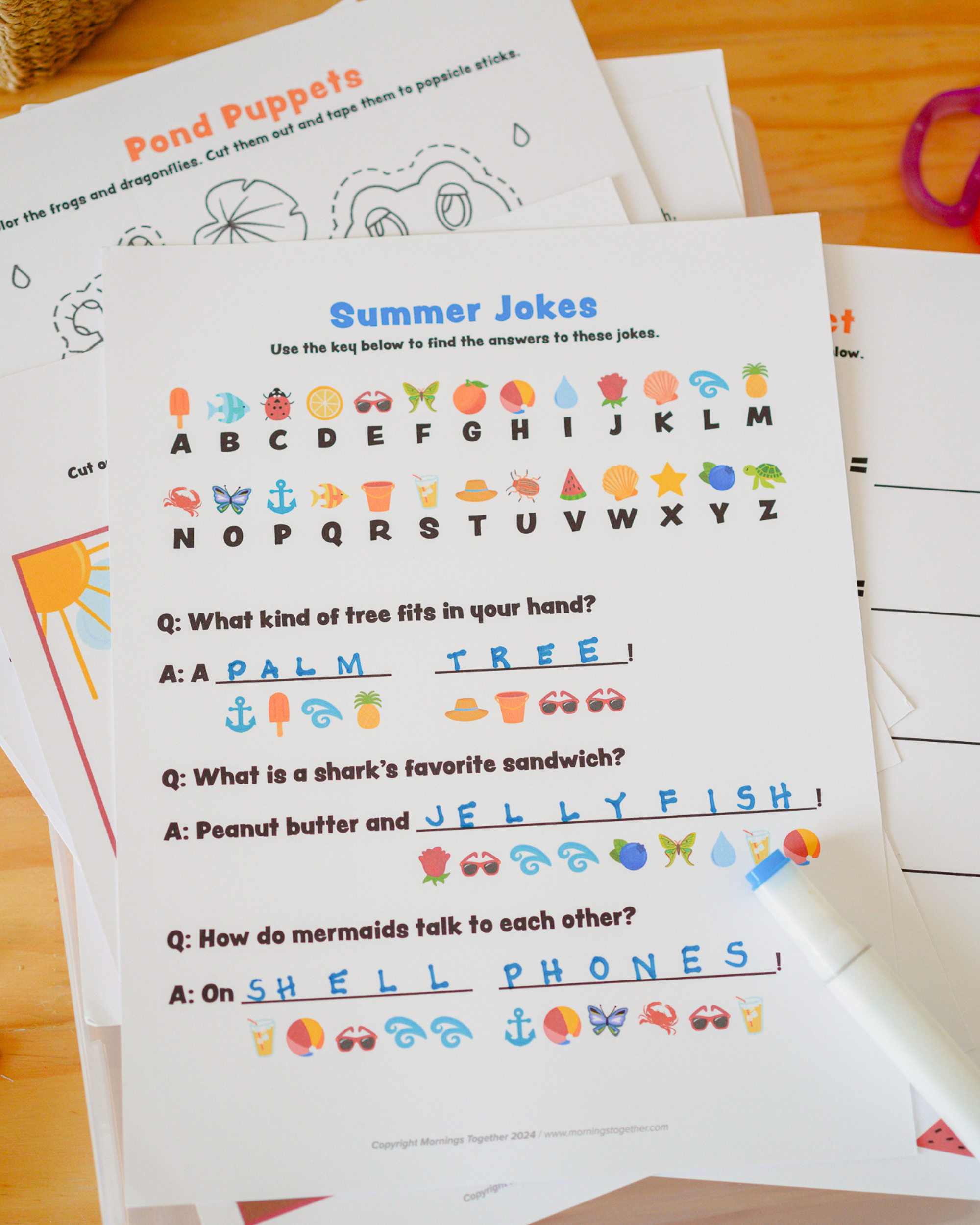A children's printable worksheet titled 'Summer Jokes' laid on top of other matching worksheets on a wooden surface. The sheet shows alphabet letters and words with small colorful summer-themed icons, plus example answers and a white marker resting on the page.