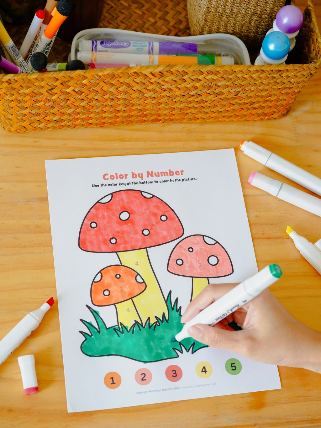 Coloring sheet with mushrooms being colored by a child using markers on a wooden table.