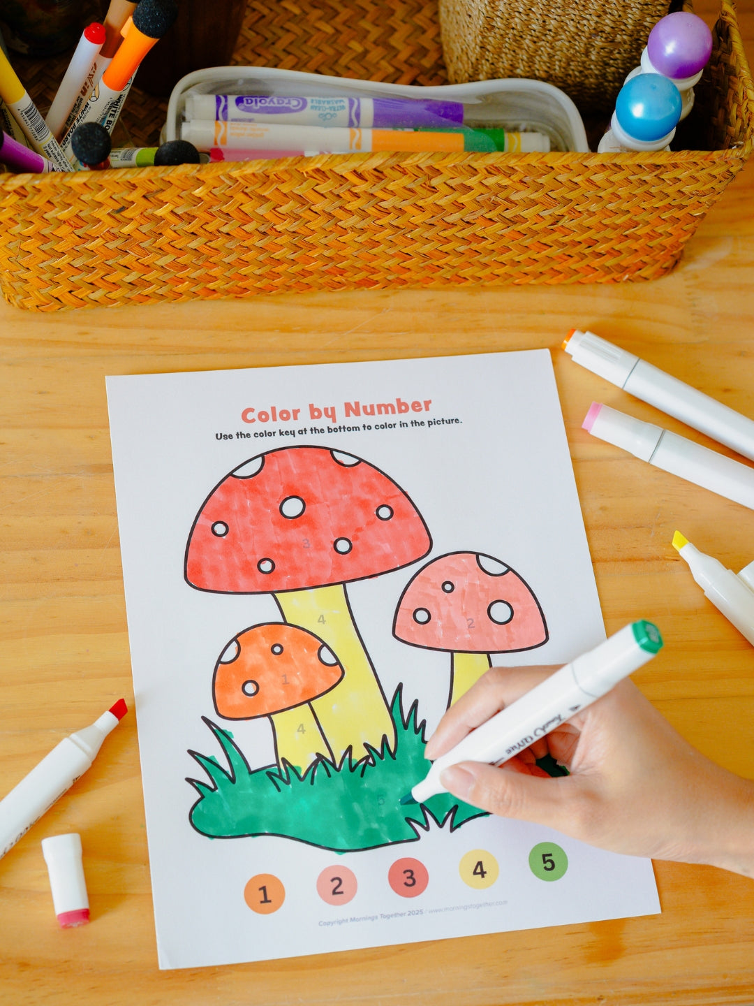 Coloring sheet with mushrooms being colored by a child using markers on a wooden table.