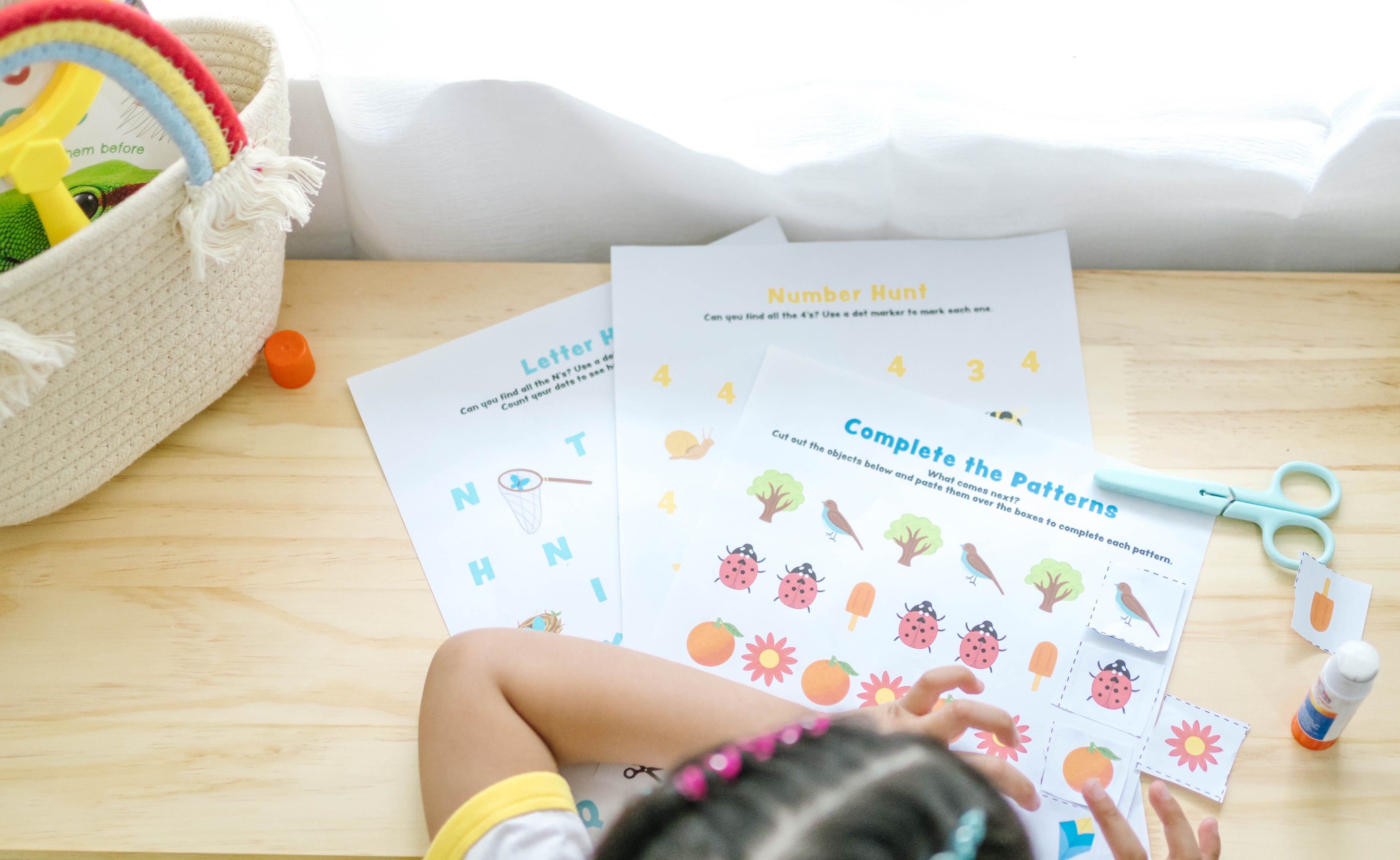 Child working on educational worksheets with a basket of toys and stationery items on a table.