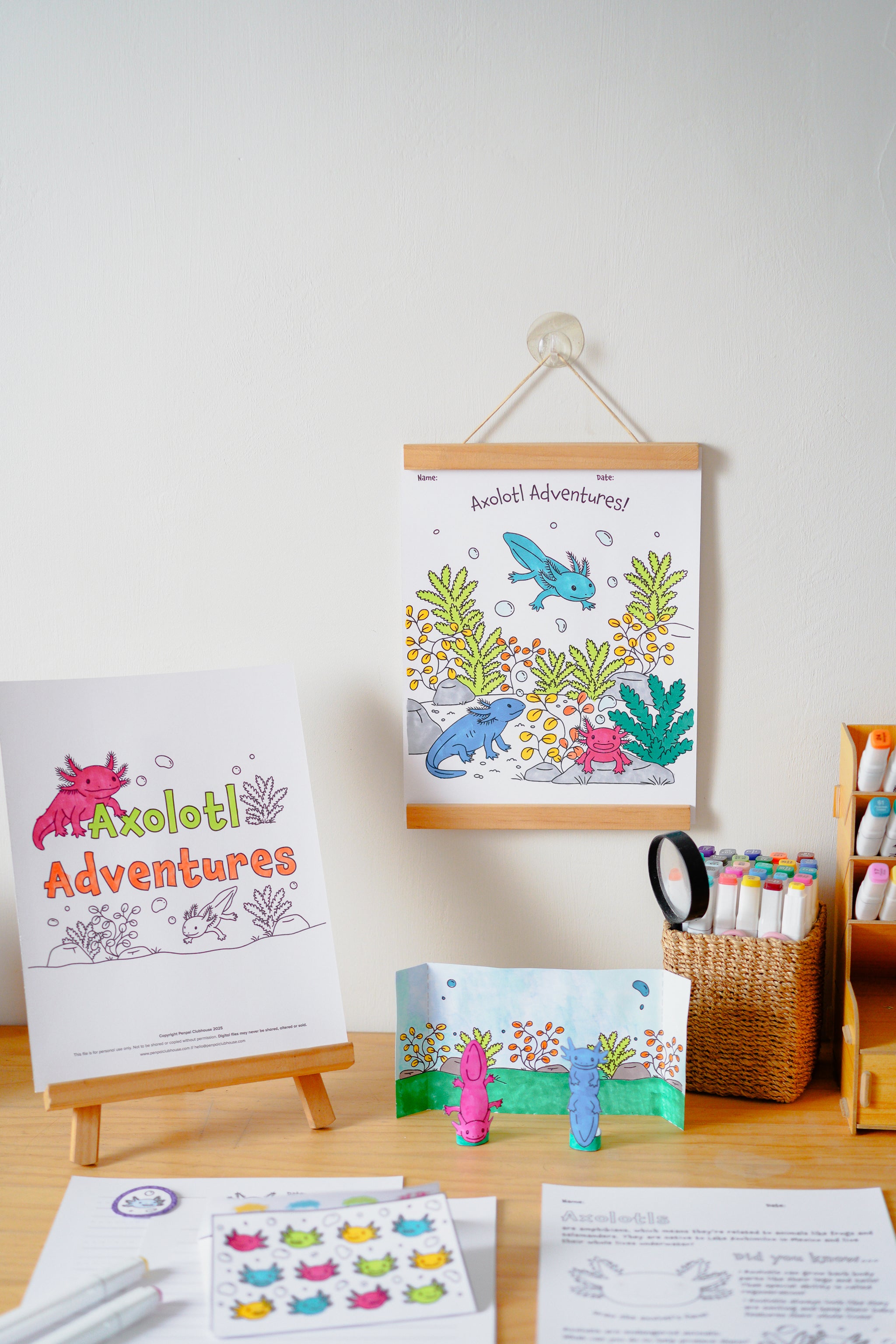 A children's art and coloring activity set displayed on a wooden desk against a white wall. The set includes a coloring book titled 'Axolotl Adventures' on a small wooden easel, a matching illustrated poster hanging on the wall, a small folded booklet, two small plastic axolotl figures, a sticker sheet, and other printed materials.