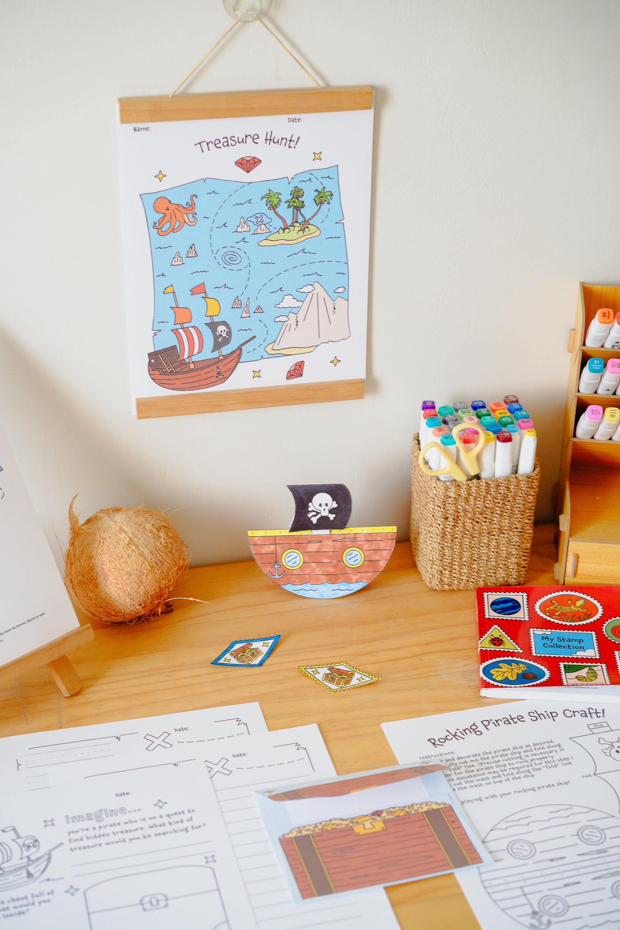 A children's pirate-themed craft and activity setup on a wooden table, featuring a hanging 'Treasure Hunt' poster, a paper ship, worksheets, a basket of markers, and a small wooden shelf with colorful items.