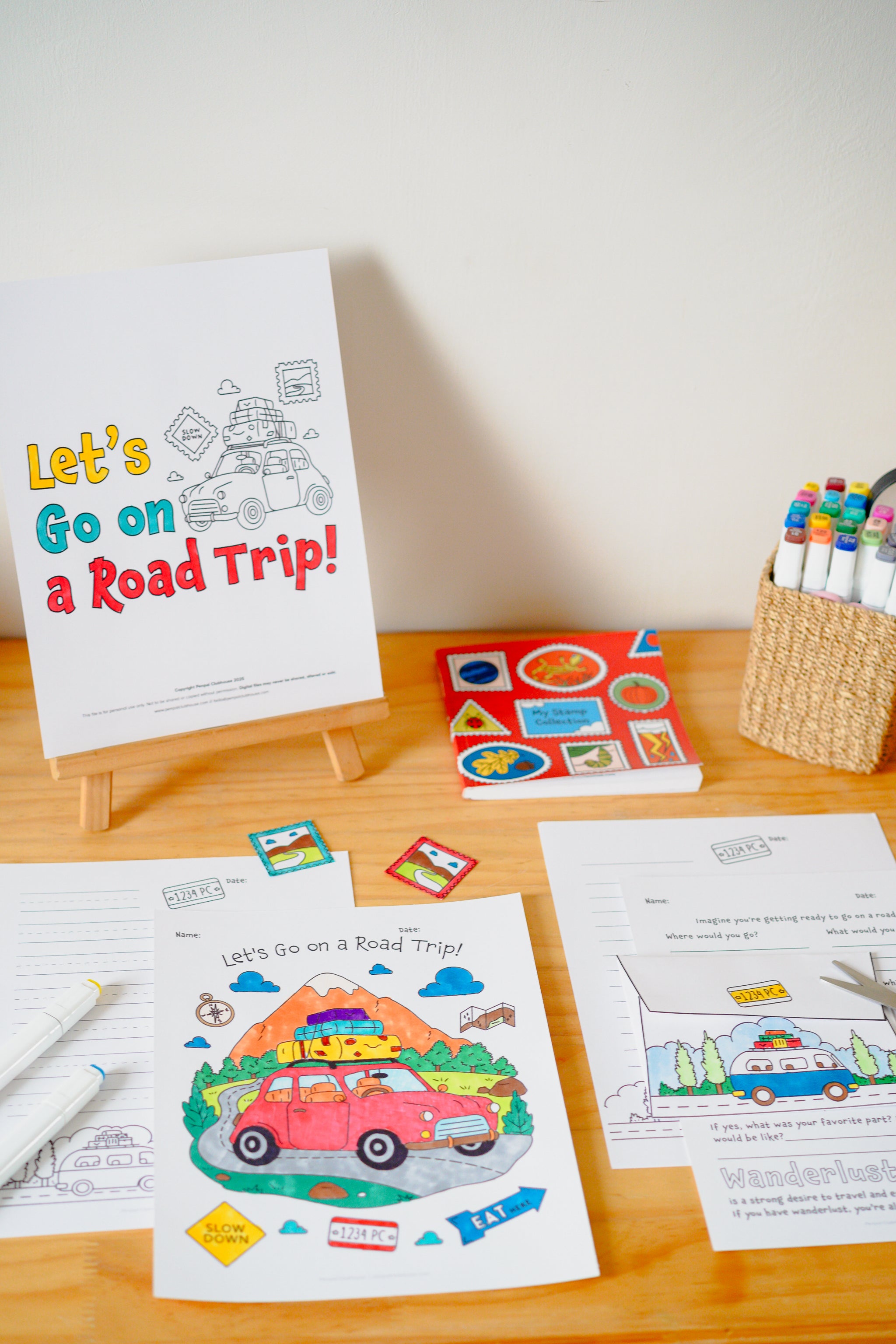 A children's road trip coloring/activity kit displayed on a wooden table against a light wall. The set includes a white cover card reading 'Let's Go on a Road Trip!', printed coloring pages, small colorful stickers or tabs, and a basket of markers.