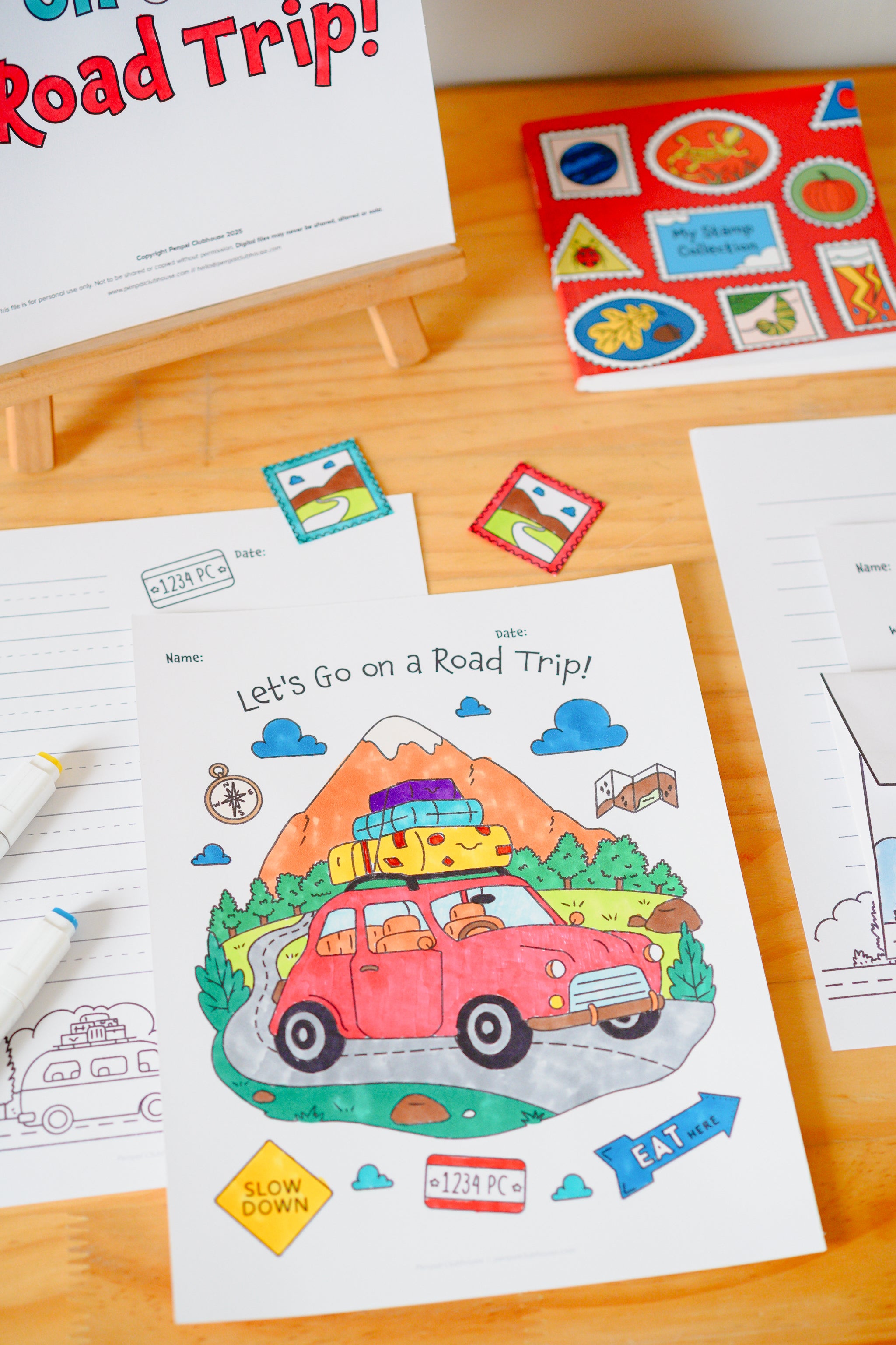 A children's road trip coloring/activity sheet on a wooden table, with a red vintage car illustration, mountains, clouds, and small travel-themed stickers around it.