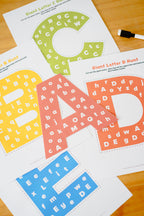 Printed letter hunt activity sheets laid out on a wooden surface, featuring oversized letters A, B, C, D, E, F, G, H, I, J, K, L, M, N, O, P, Q, R, S, T, U, V, W, X, Y, Z with hidden alphabet words inside. A small black marker is visible at the top right.