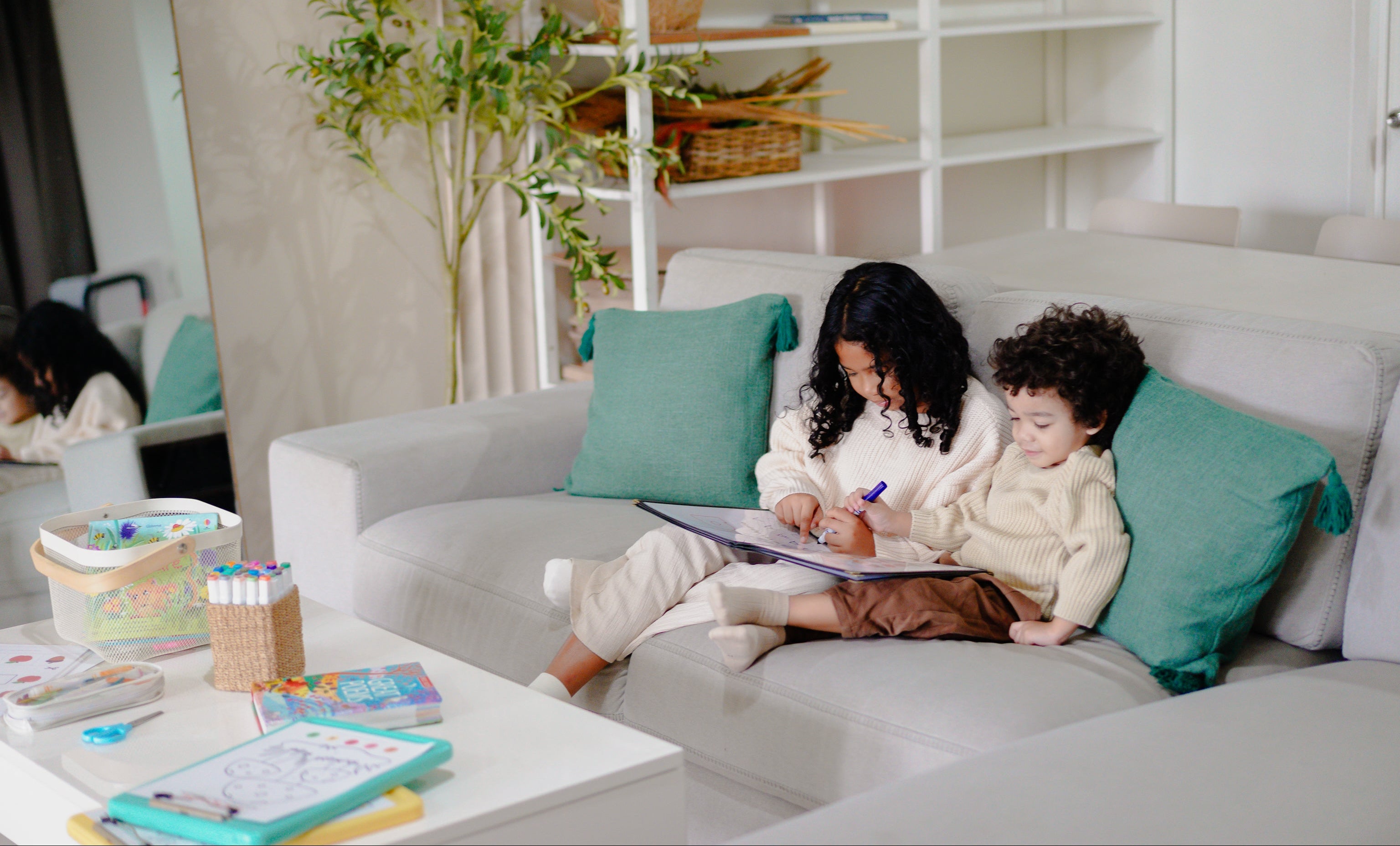 Two children sitting on a couch in a living room with art supplies.