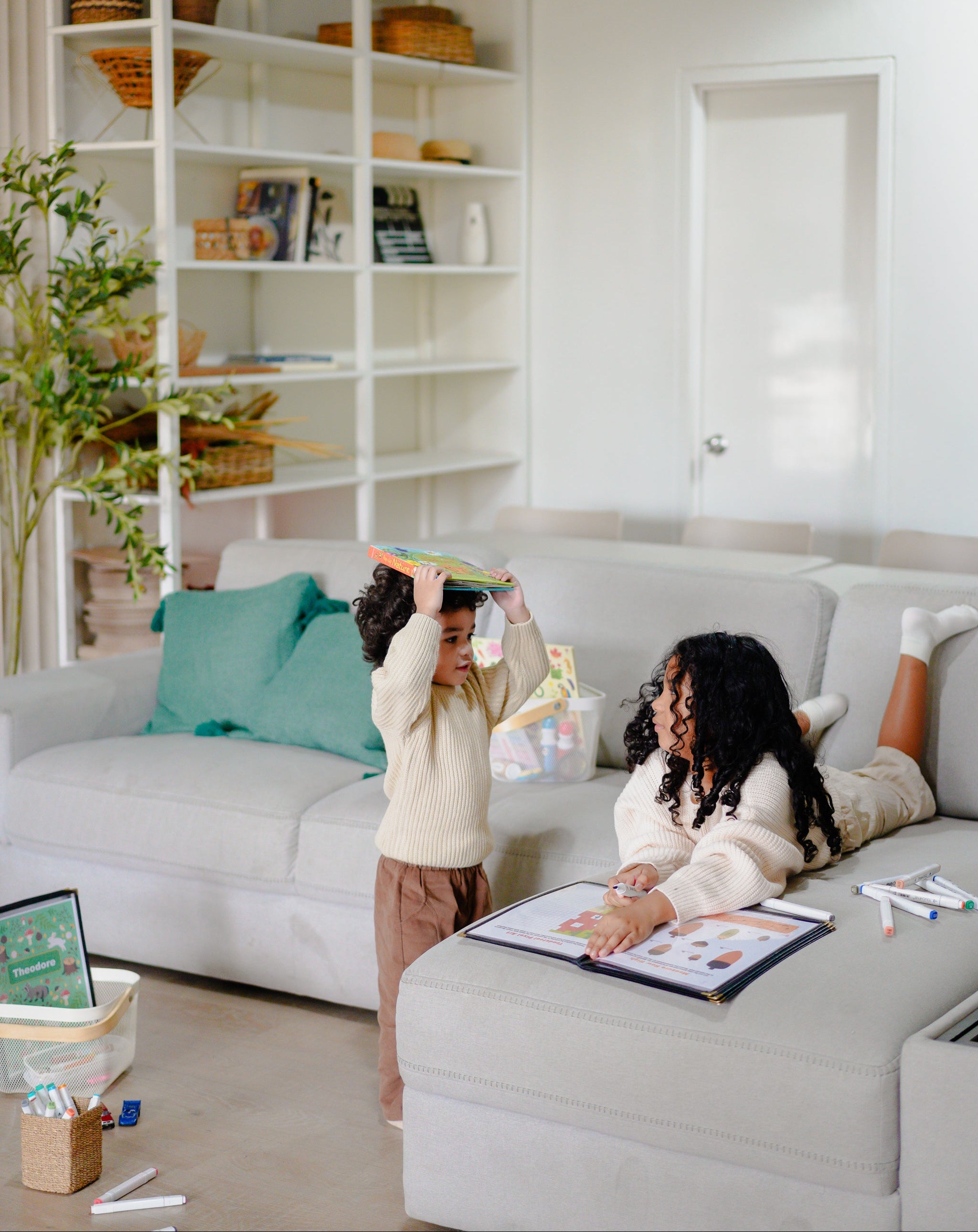 Two children playing on a gray sofa in a living room with a white bookshelf and decor.