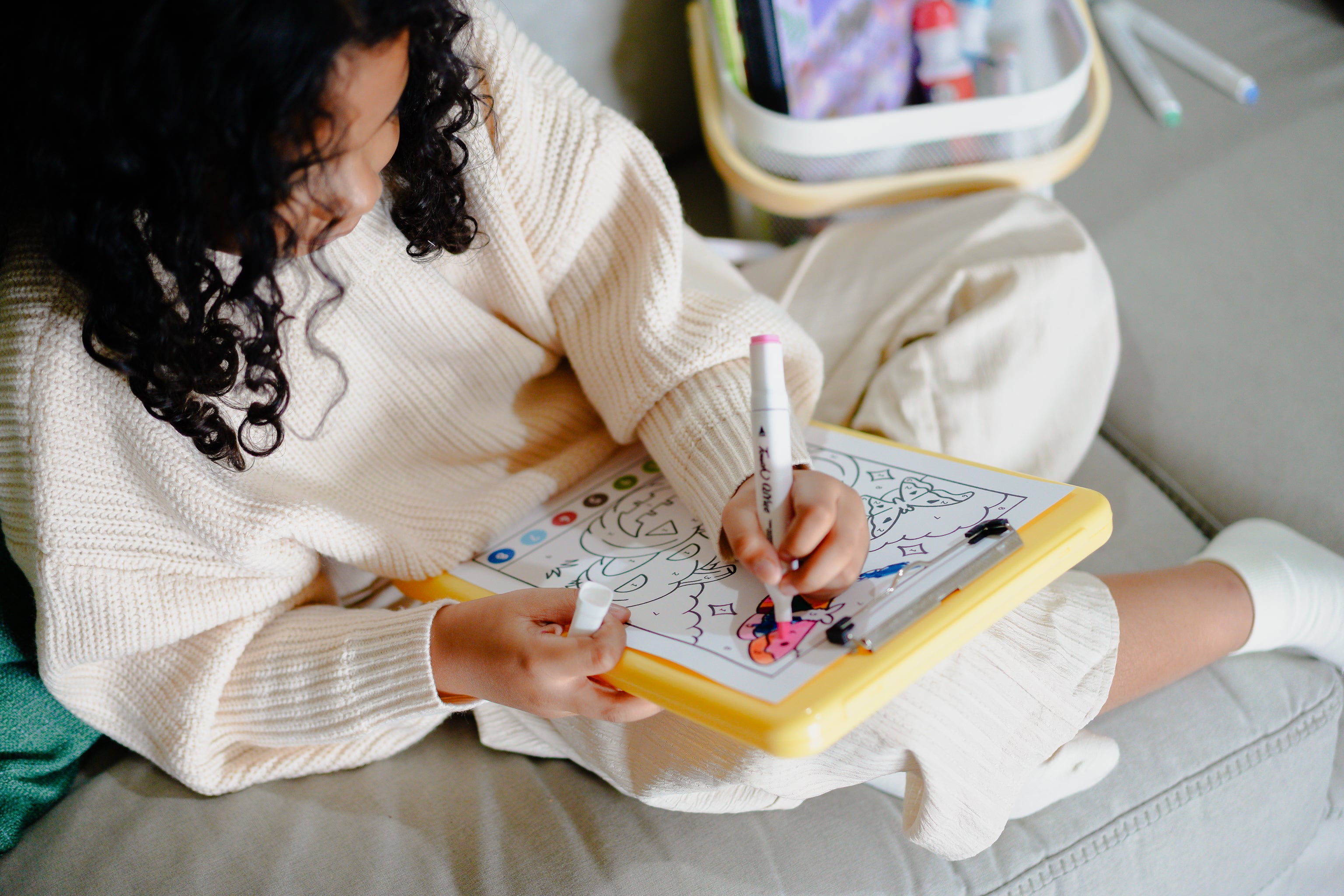 Young girl coloring on coloring page on a yellow clipboard, with markers while sitting cross-legged on a couch