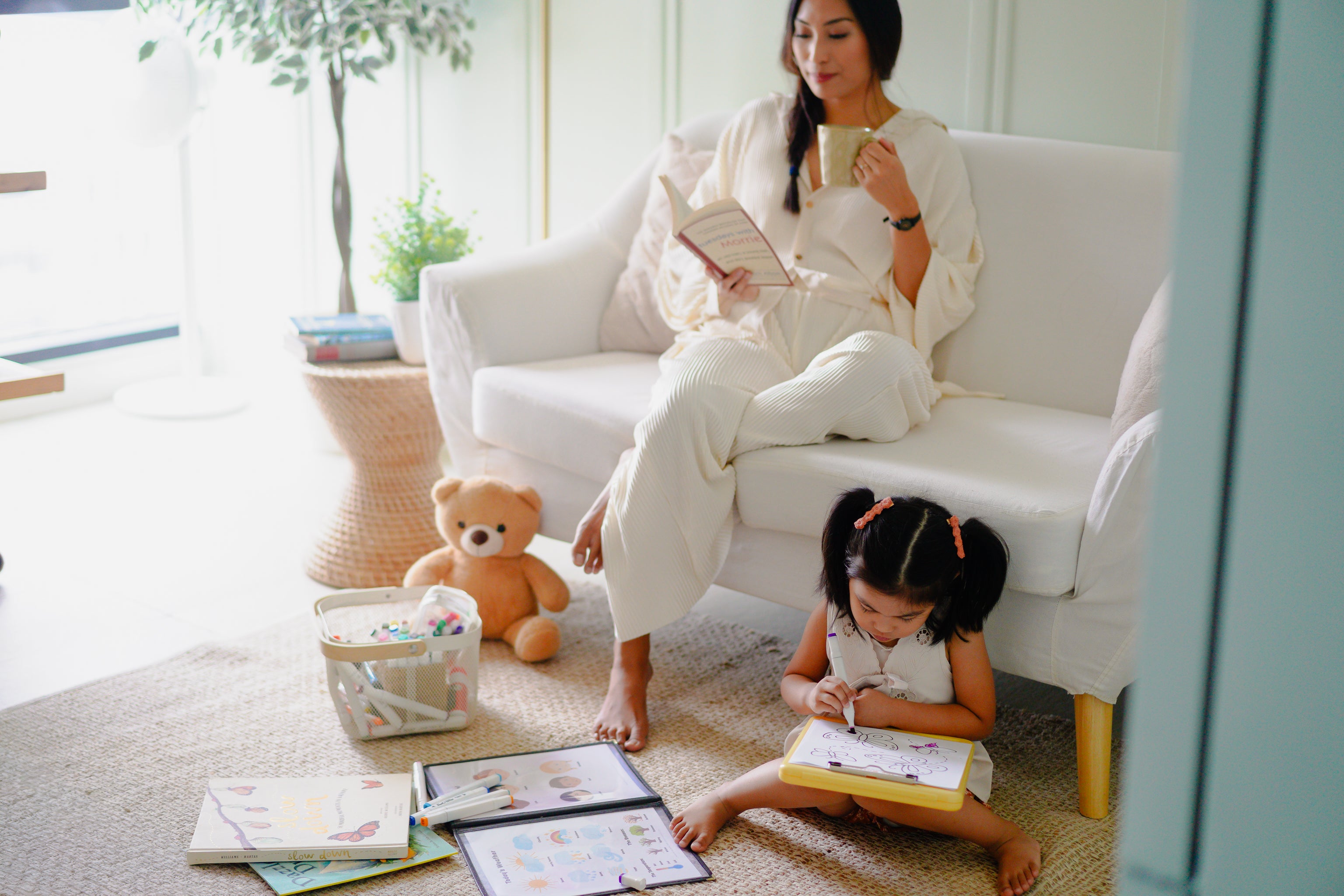 Woman reading a book to a child on a white couch in a bright living room.