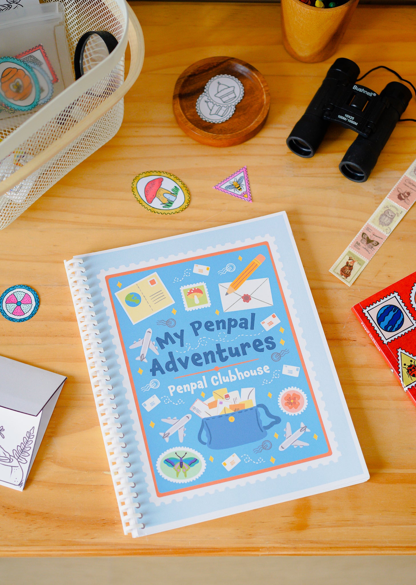 A spiral-bound notebook titled 'My Penpal Adventures' by 'Penpal Clubhouse' lies on a wooden desk surrounded by travel-themed stickers, binoculars, and other stationery items.