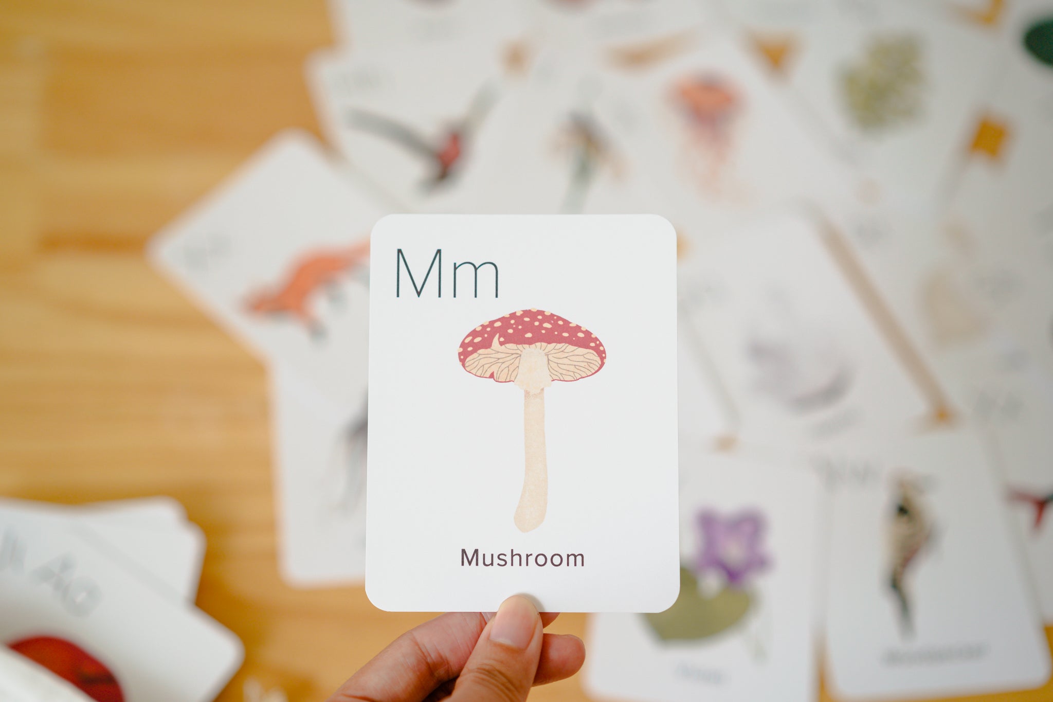 A hand holds a white educational card featuring a mushroom illustration and the letter combination 'Mm' over a wooden table, with other illustrated cards blurred in the background.