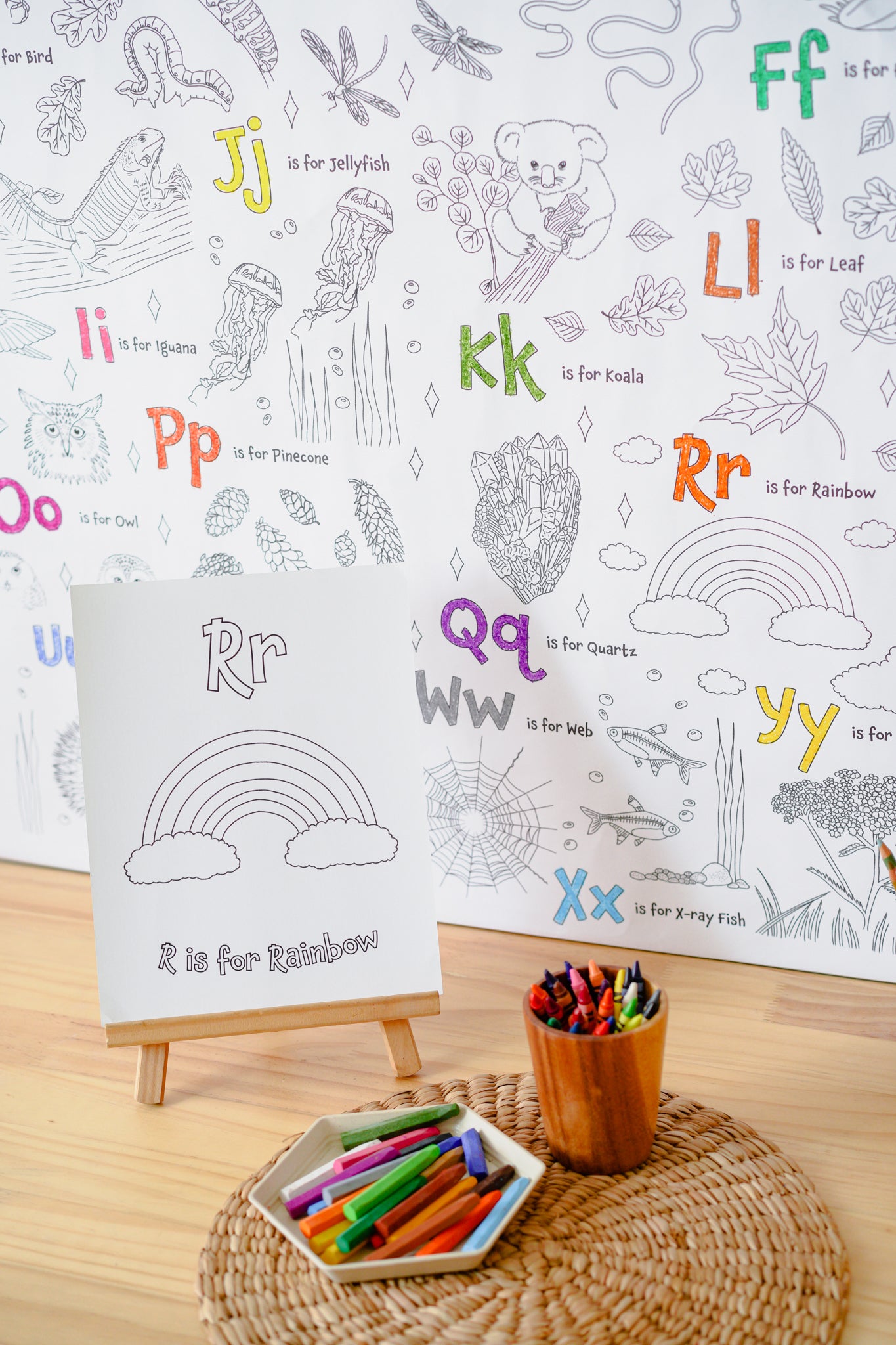 A children's alphabet coloring/activity book page for the letter 'Rr' with a rainbow illustration, displayed on a small wooden easel. The scene includes a cup of colored pencils and a tray of crayons on a woven mat, with a larger matching alphabet coloring sheet in the background.
