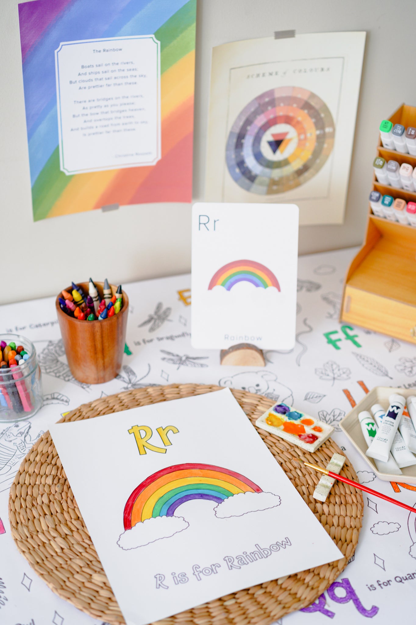 A children's alphabet learning setup featuring a card and print with the letter 'Rr' and a rainbow illustration, displayed on a table with art supplies, crayons, paint tubes, and educational posters.