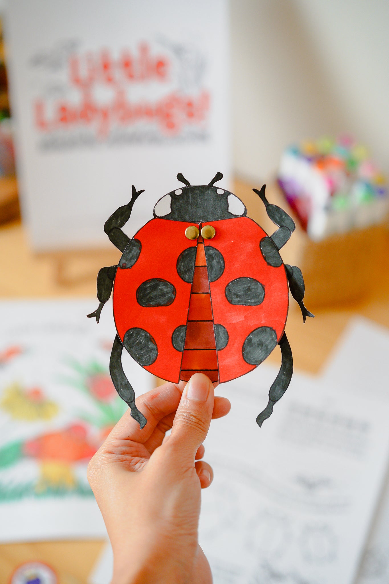 ladybug paper craft being held up by a hand in front of educational printables blurred out in the background.