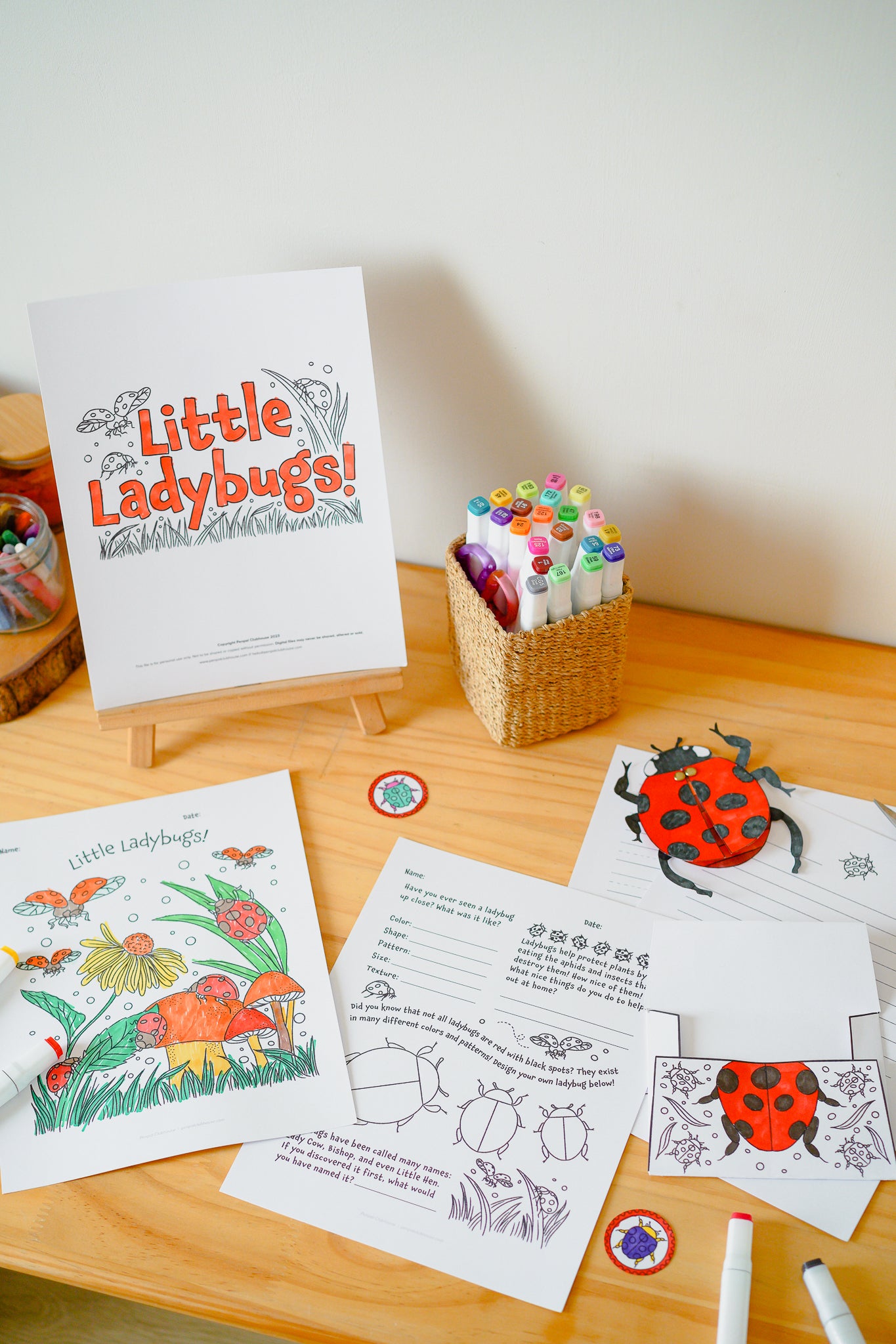 A children's ladybug-themed activity set arranged on a wooden table against a light wall. The set includes a printed sign reading 'Little Ladybugs!', coloring pages, printed worksheets, ladybug craft cutouts, a small woven basket holding markers, and a few colored markers.