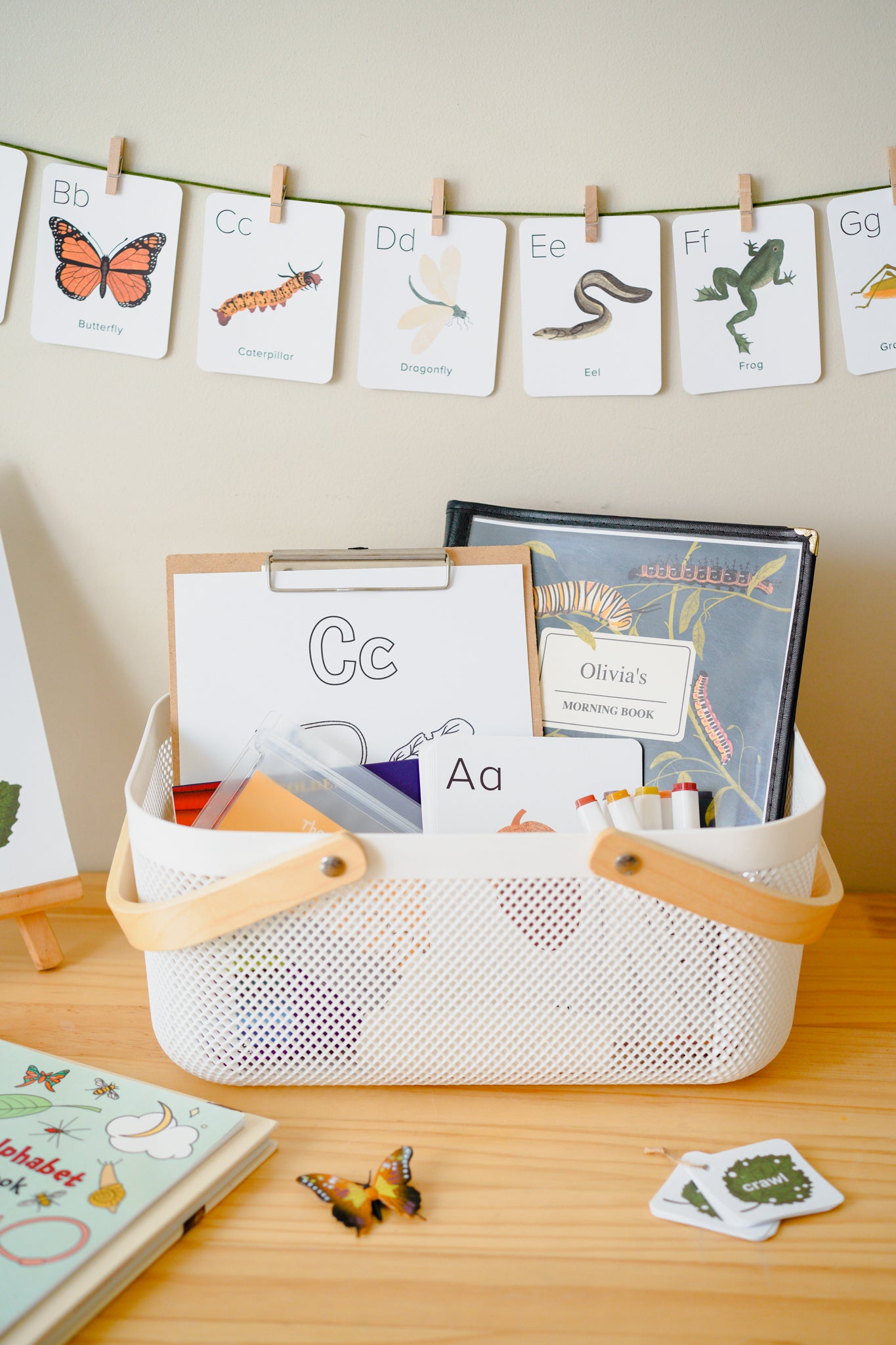 A white mesh storage basket with light wood handles sits on a wooden table, filled with alphabet cards, a clipboard, and a book. A matching alphabet banner hangs on the wall behind it.