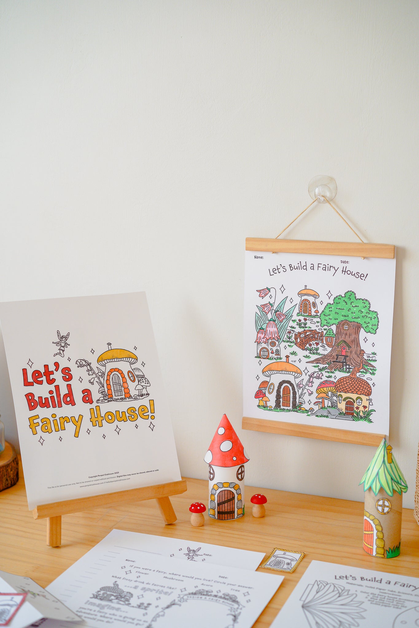 A children's fairy house coloring/activity set displayed on a light wood table against a white wall, including a printed sign reading 'Let's Build a Fairy House!', a framed illustrated poster, small toy houses, and printed sheets.