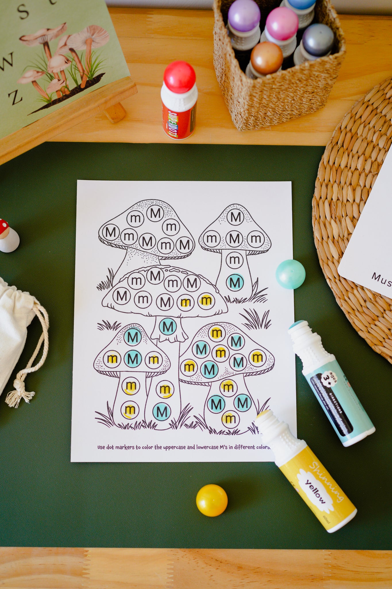 A children's coloring activity sheet featuring mushroom outlines with letters inside, placed on a dark green mat with craft supplies around it, including paint bottles, a wicker basket, a small drawstring pouch, and two small balls.