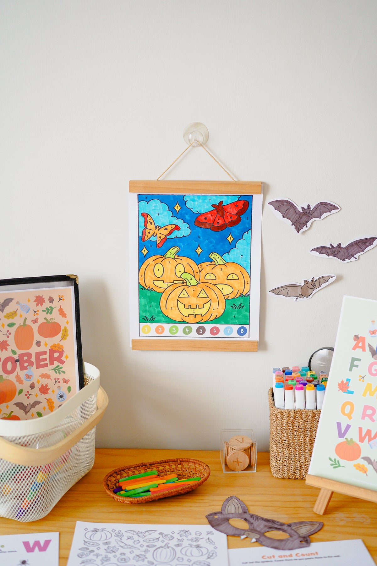 A colorful children's wall hanging poster featuring jack-o'-lanterns, a red fish, and a rainbow, displayed on a wooden table with art supplies, stickers, and other educational items.