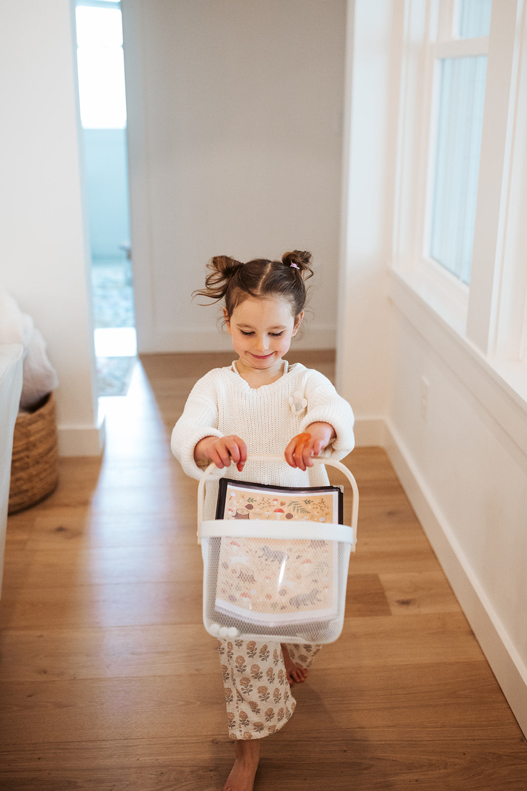 Child holding a basket with items on a wooden floor in a bright room.