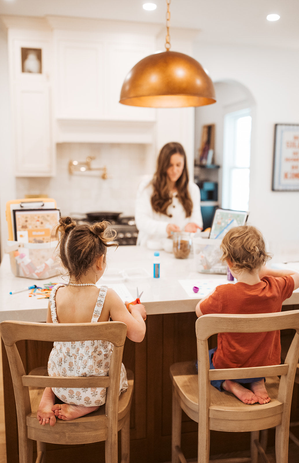 Two children sitting at a kitchen counter with a woman in the background.